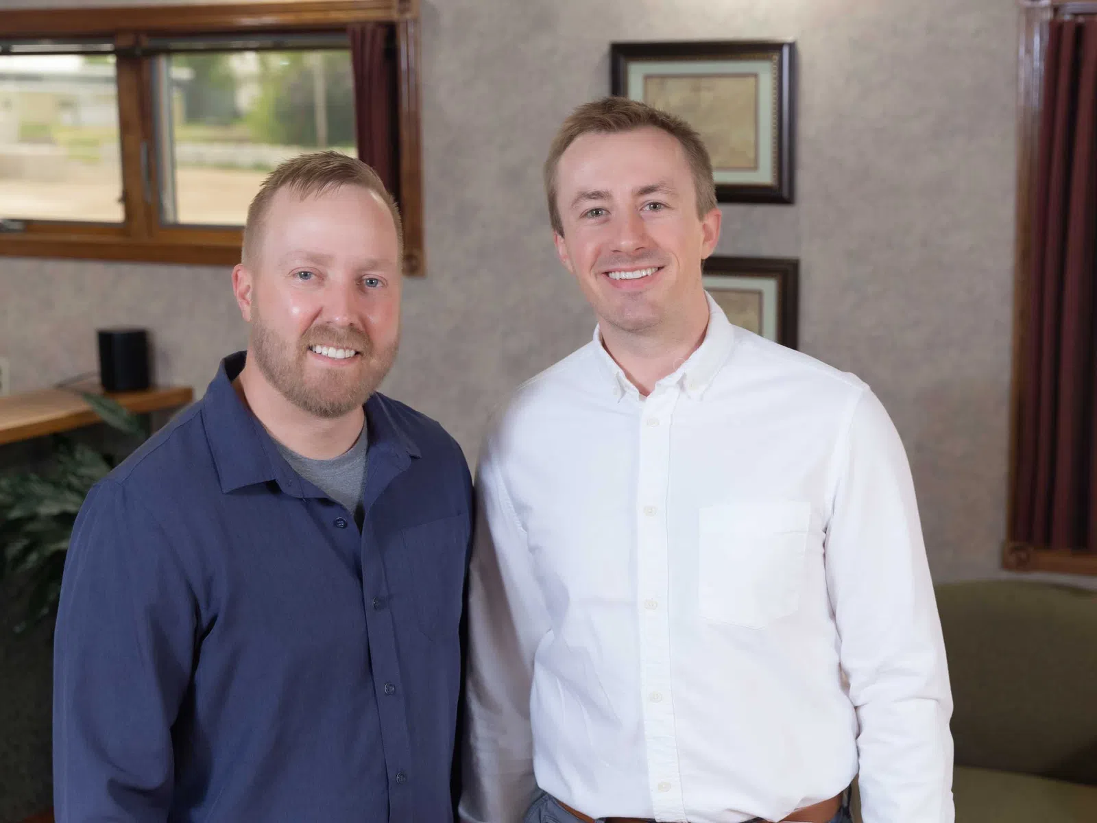 Two smiling men in business casual shirts posing indoors with framed pictures and windows in the background