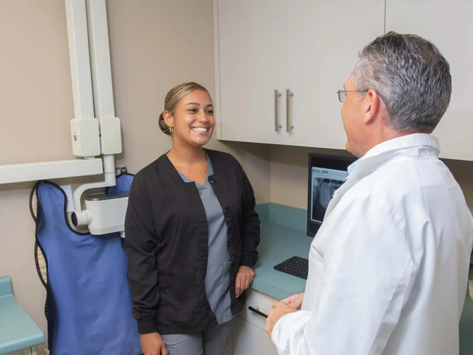 Smiling female dental assistant talks with male dentist in clinic with X-ray visible on computer screen