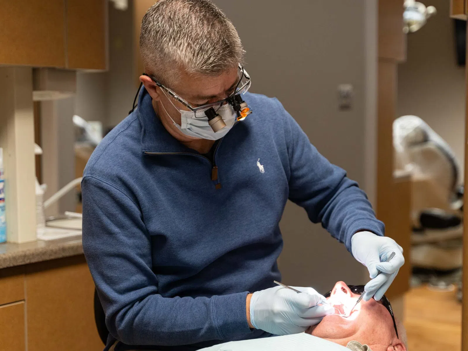 Dentist wearing magnifying glasses and gloves examining a patient's teeth in a dental clinic.