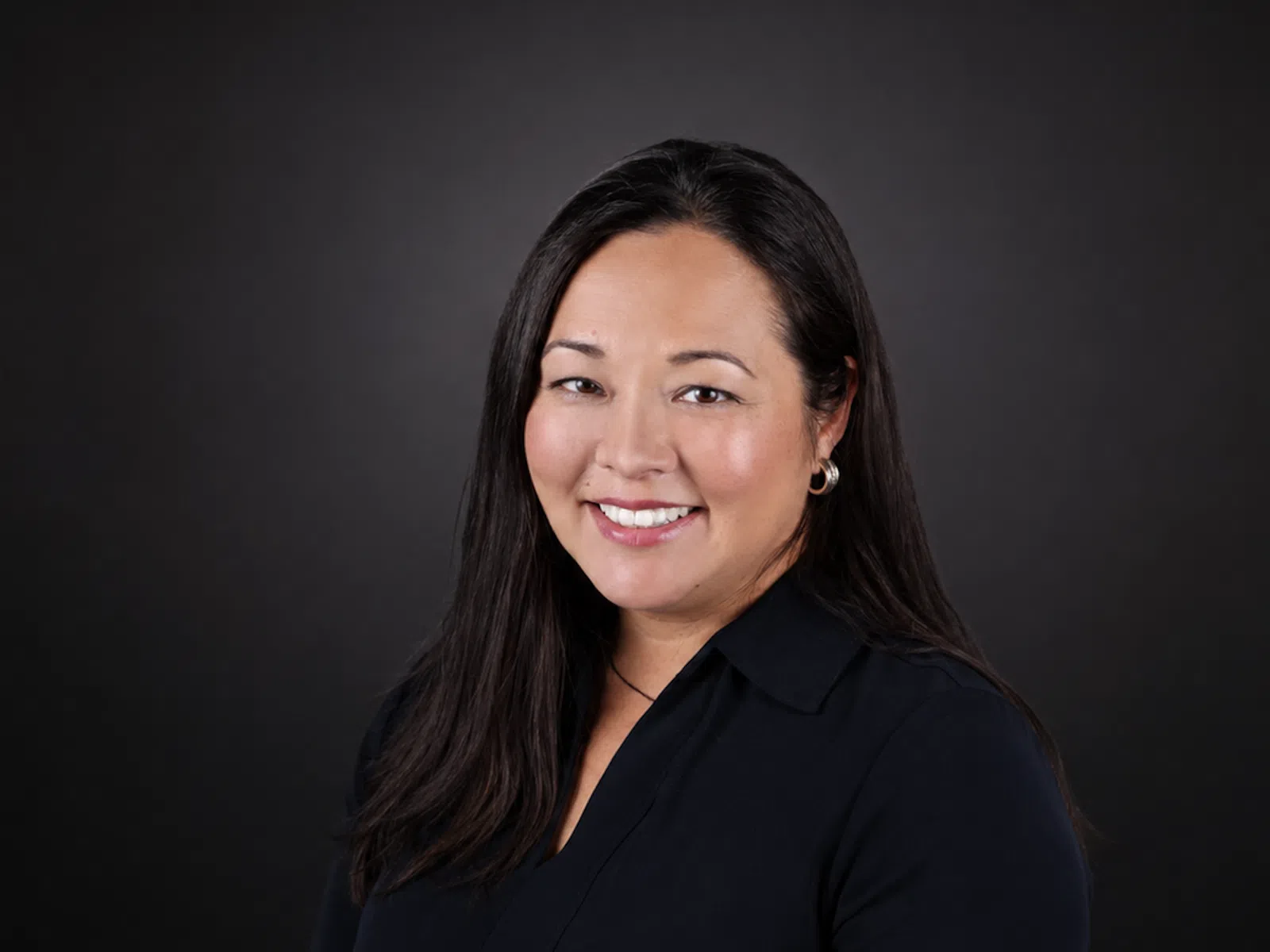Smiling woman with long black hair wearing a dark blouse and silver hoop earrings against a black background