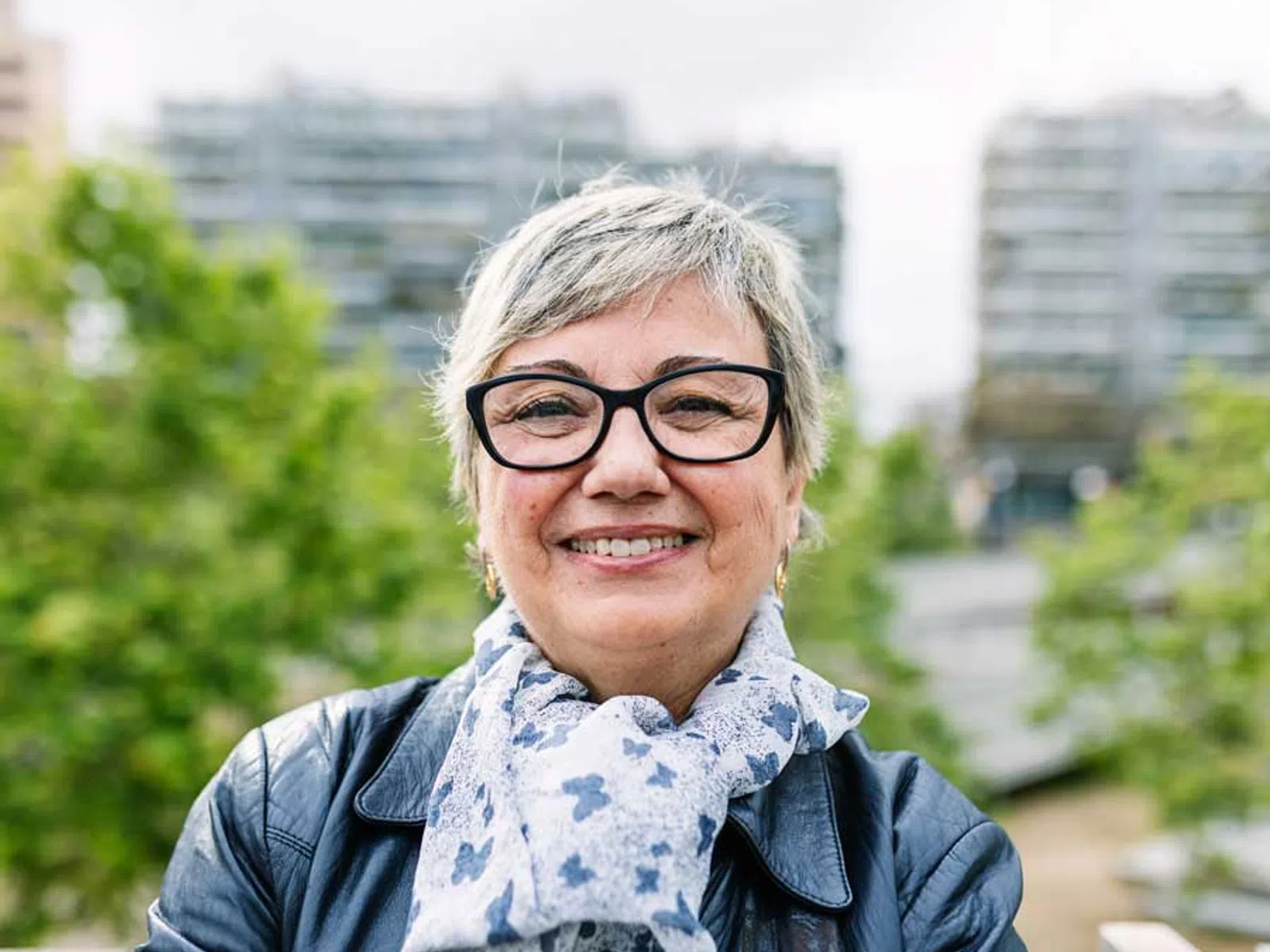 Smiling older woman with glasses wearing a patterned scarf and black leather jacket outdoors in urban park.