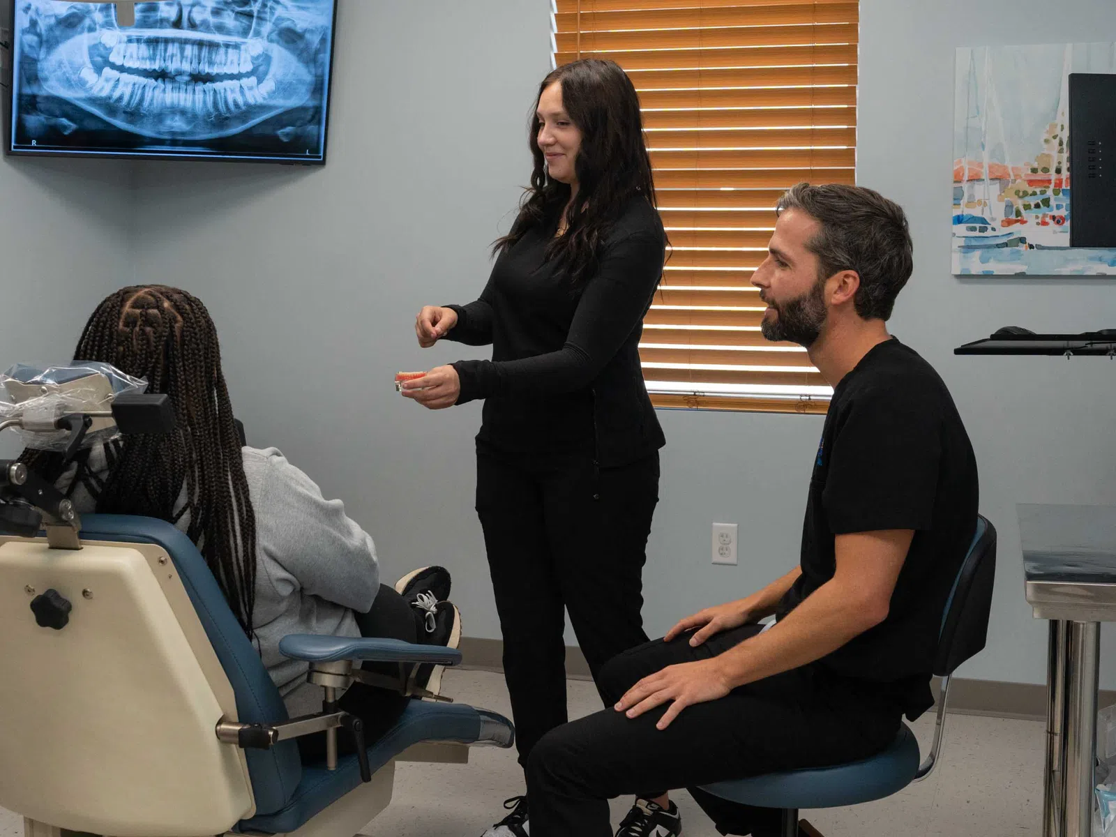 Dentist and assistant consulting patient seated in dental chair with tooth x-ray on screen.