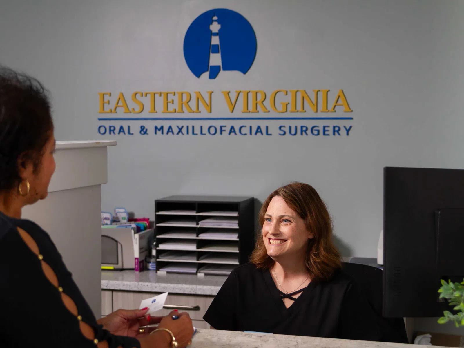 Receptionist smiling at patient in office with Eastern Virginia Oral & Maxillofacial Surgery signage with overlaid text.