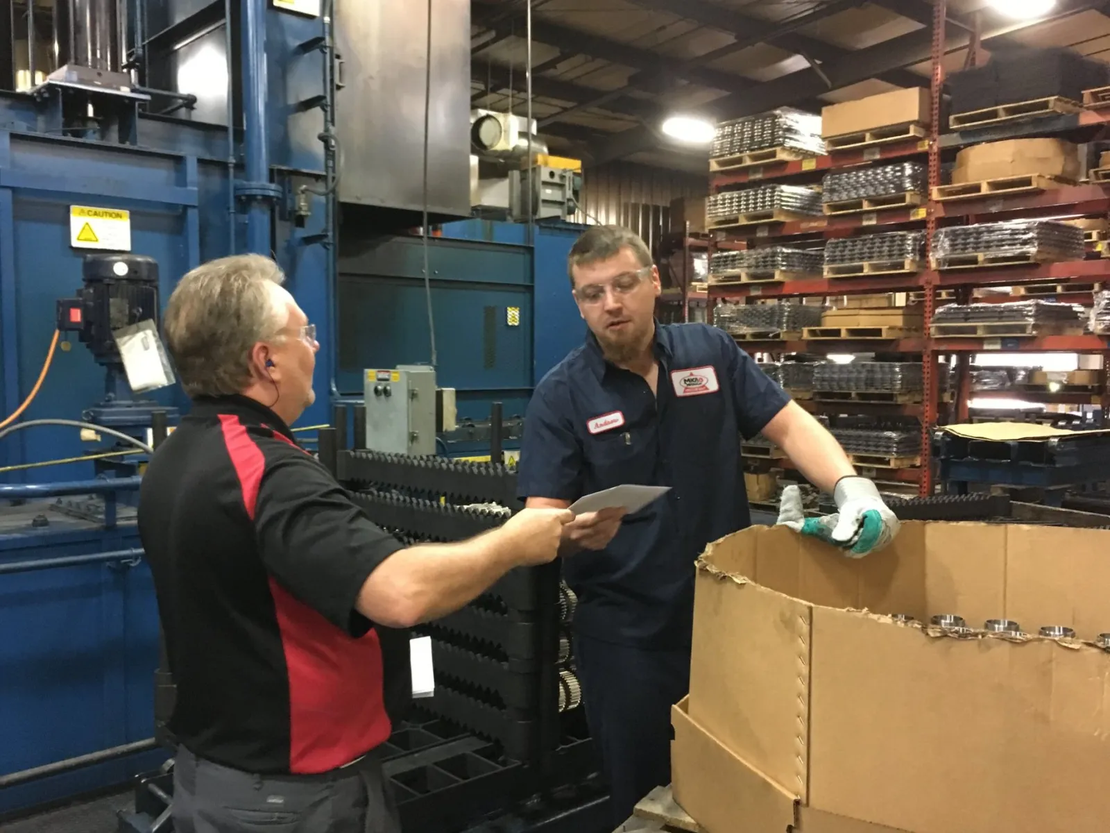 Two factory workers exchanging documents in an industrial warehouse near machinery and shelves with metal parts.