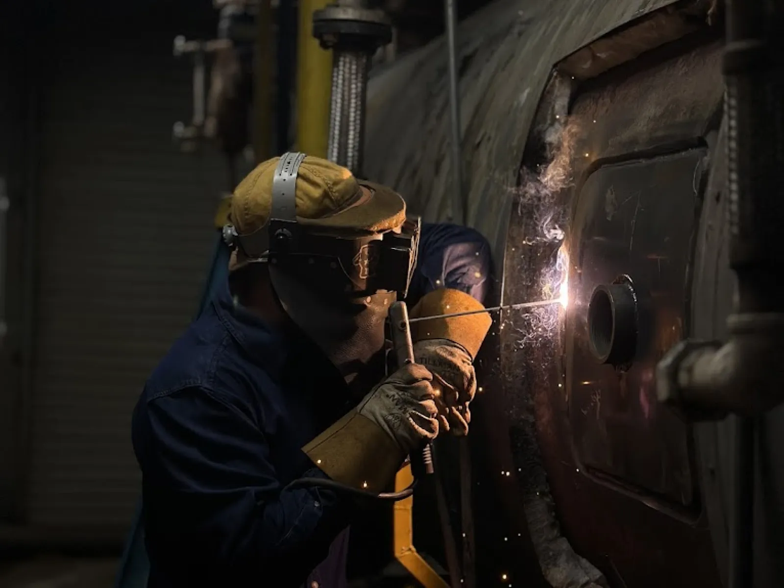 Worker wearing protective gear welding a large industrial metal tank, sparks flying in dim workshop light