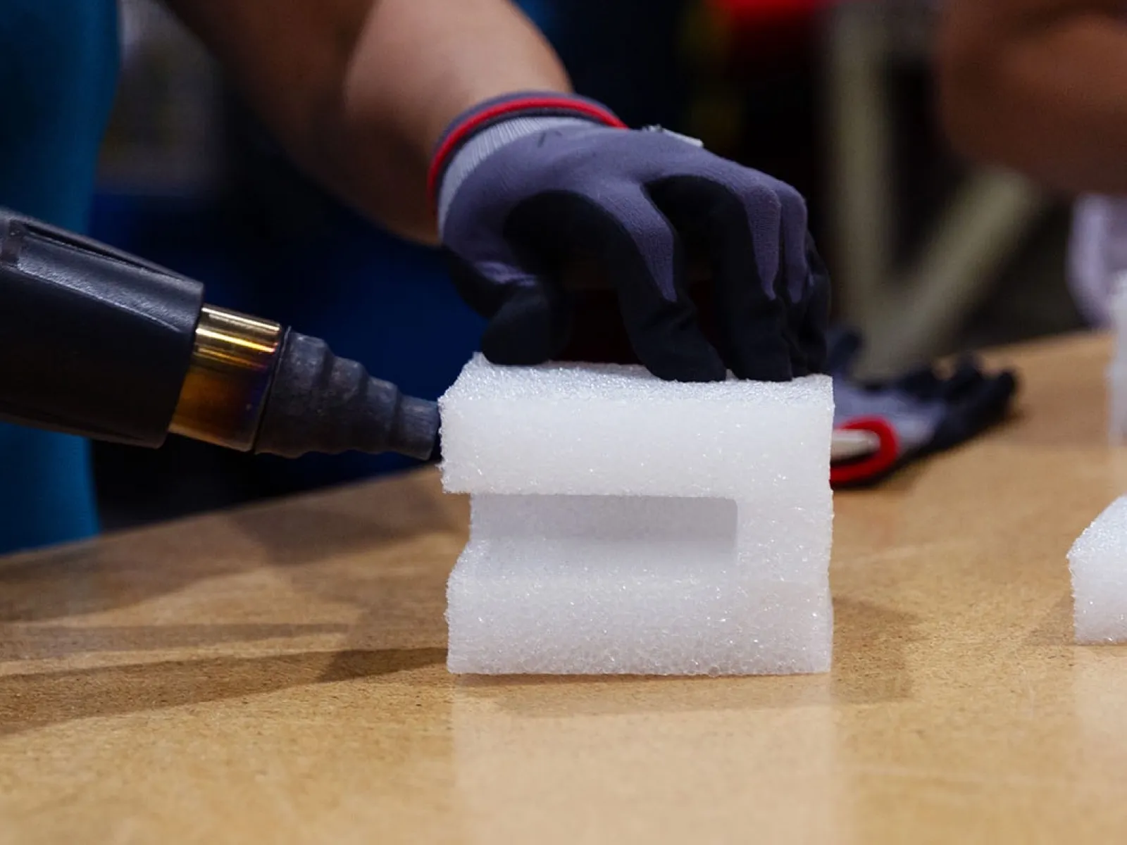 Person wearing gloves using a heat gun to shape white foam block on a wooden table.