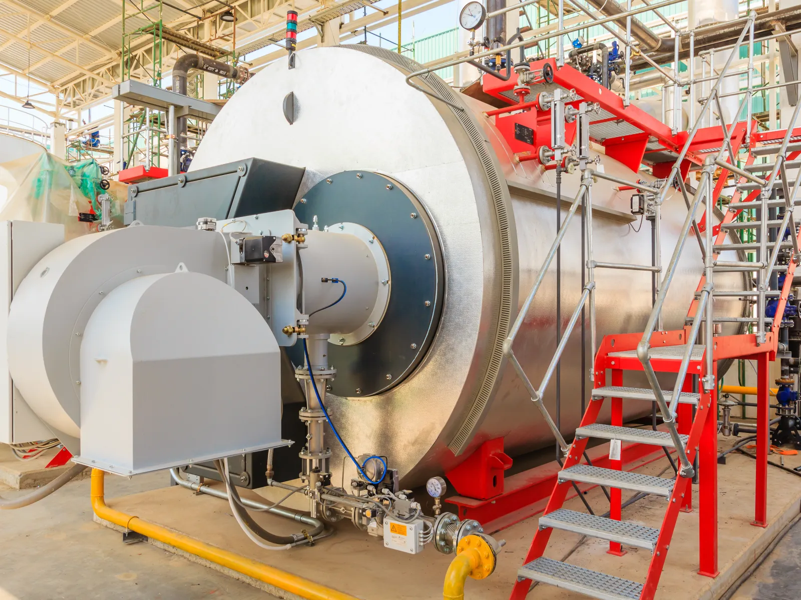 Large industrial boiler system with metal pipes and red safety stairs in a factory setting.