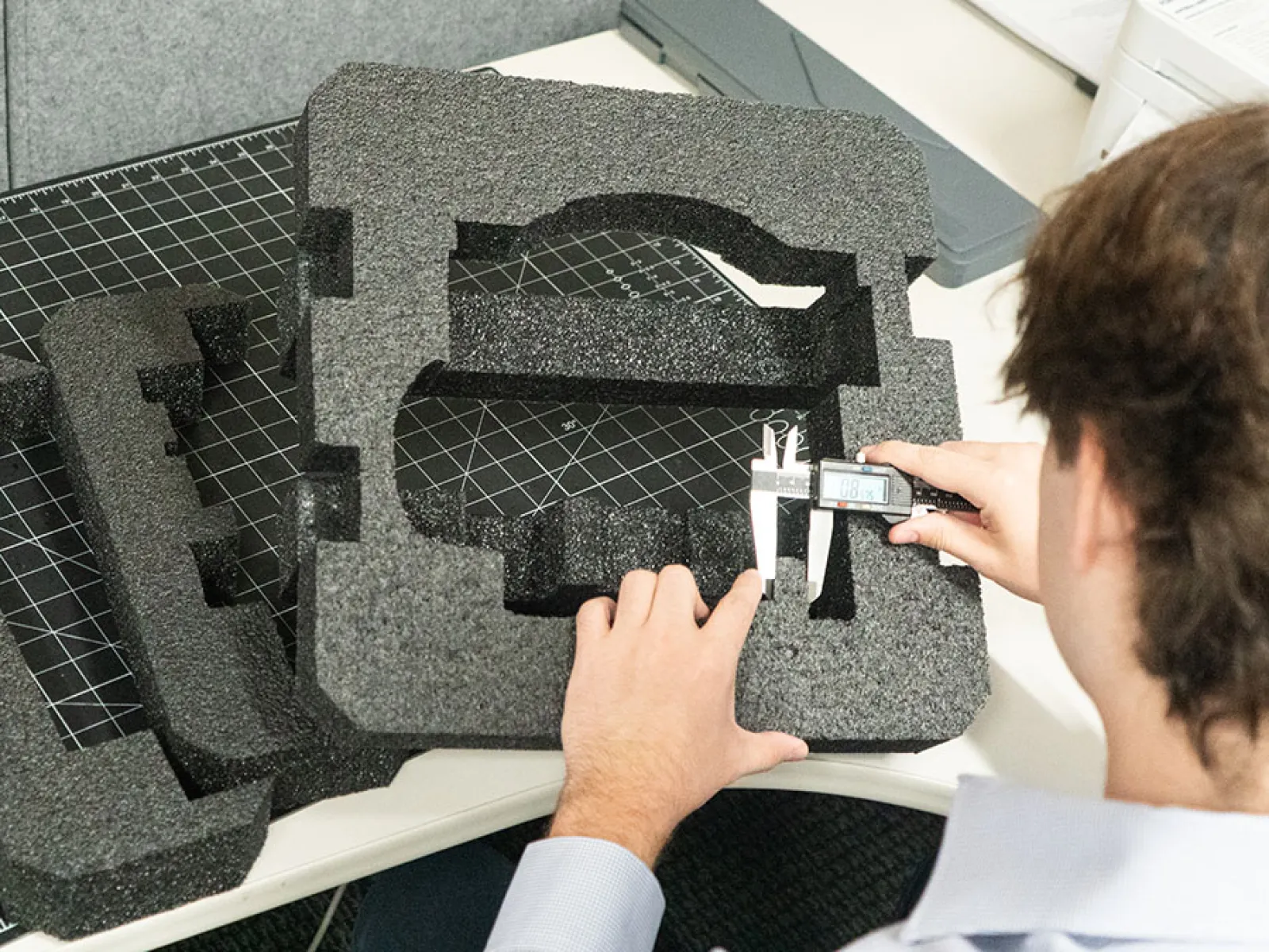 Person measuring custom foam packaging insert with caliper on a workbench with cutting mat.