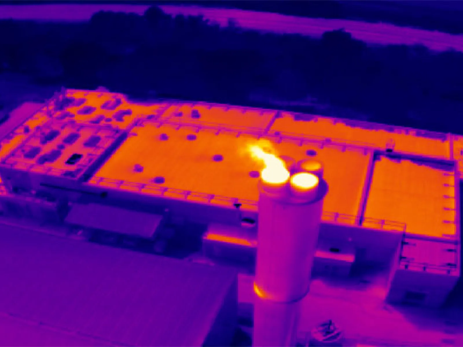 Thermal image of factory rooftop and smoke stack emitting heat visible as bright spots against cooler background.