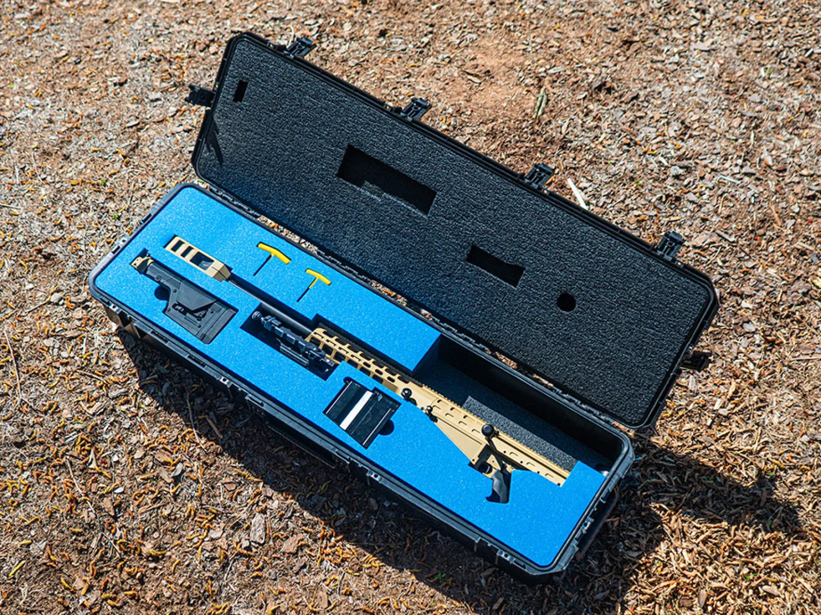 Tactical rifle with accessories neatly placed inside a blue foam-lined black hard case on the ground outdoors.