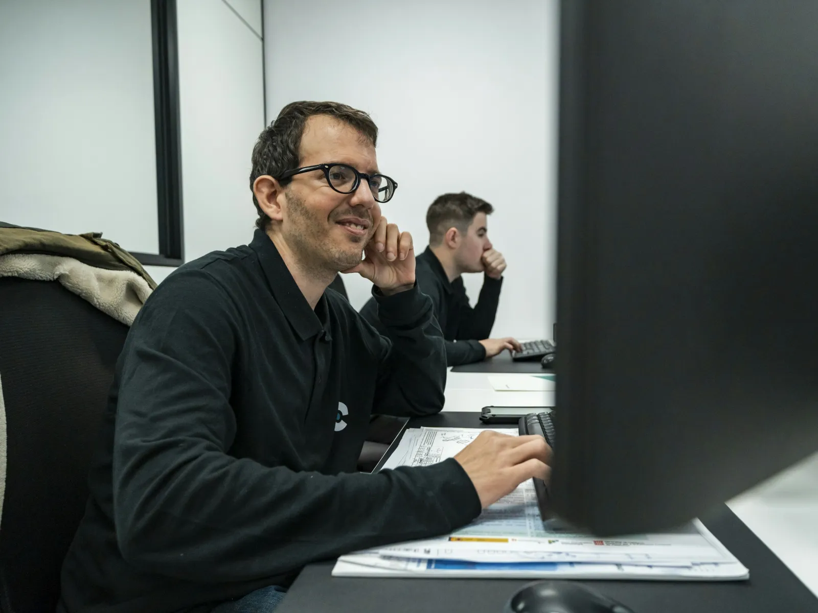 Two men working at computers in an office with documents and focused expressions.
