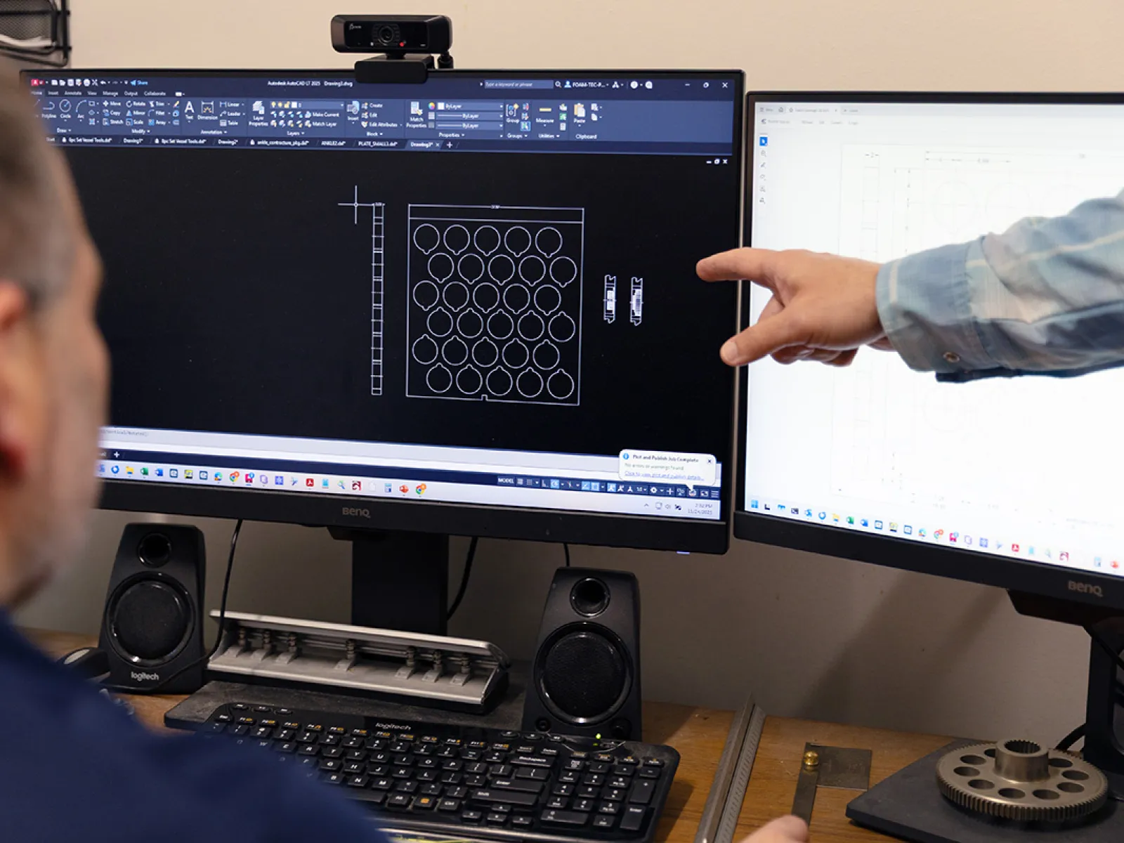 Engineer reviewing CAD design of mechanical components on dual monitors with keyboard and speakers on desk.