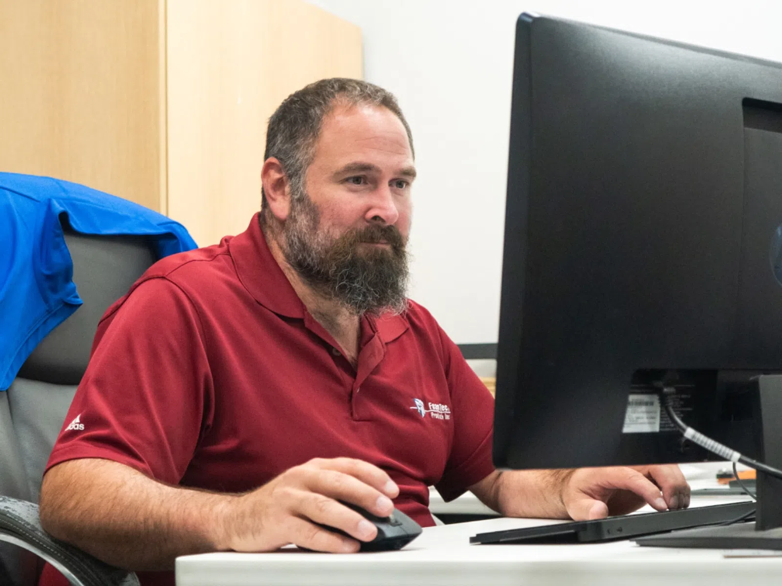 Man in red polo shirt working on desktop computer at office desk with blue jacket on chair