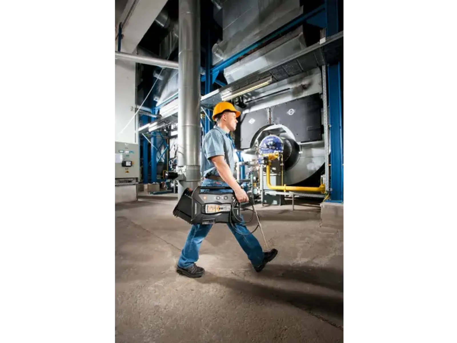 Industrial technician in safety gear walking with equipment inside a factory or plant environment.
