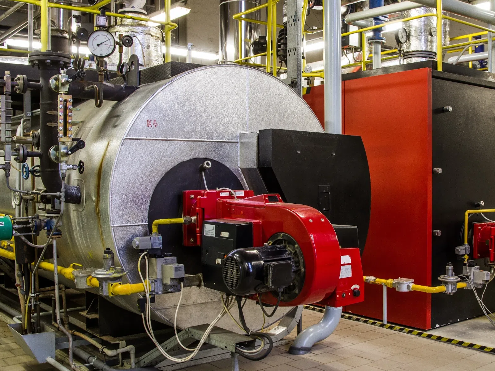 Industrial boiler system with red fuel burner, pressure gauges, pipes, and control valves in a factory setting