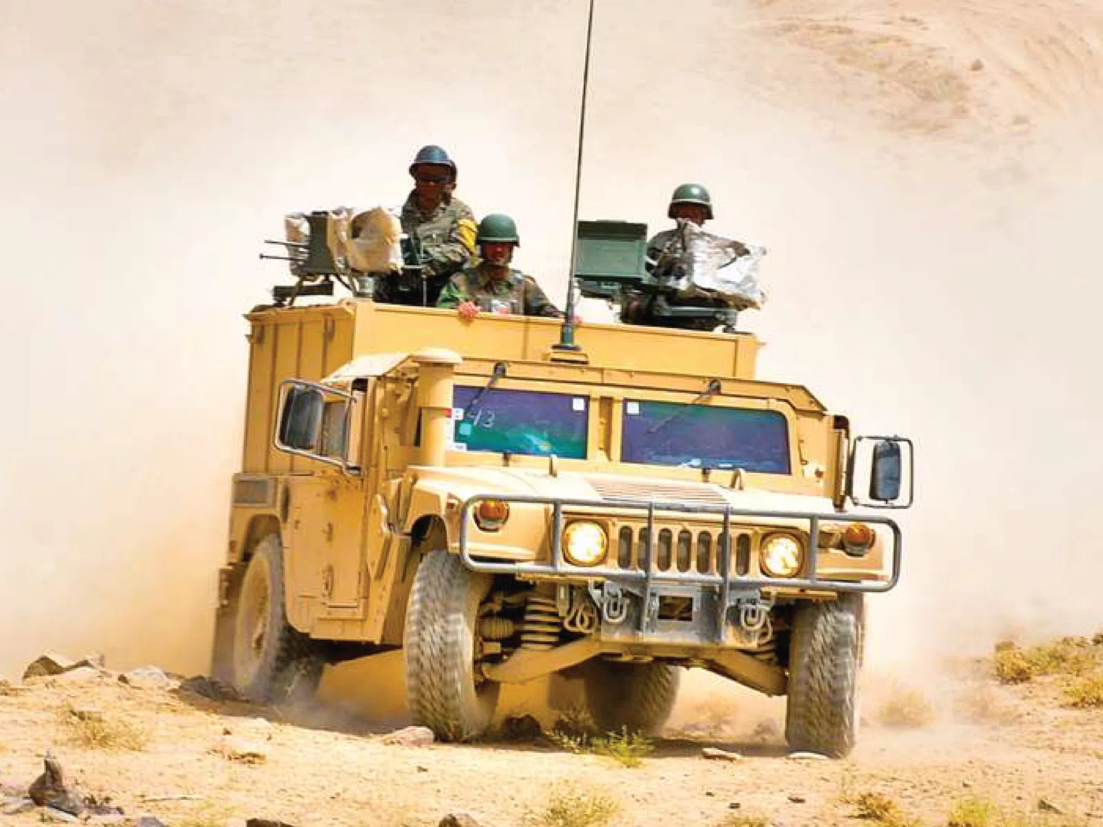 Military Humvee with armed soldiers driving on dusty desert terrain under bright daylight.