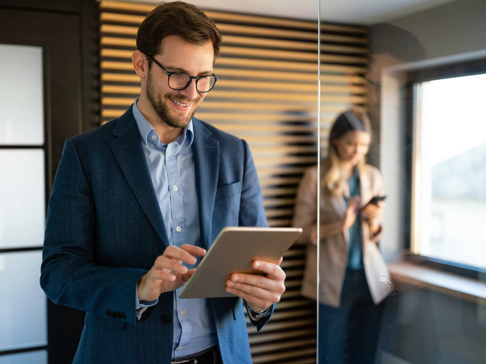 Smiling businessman using a tablet while a woman checks her phone in a modern office with wooden accents.