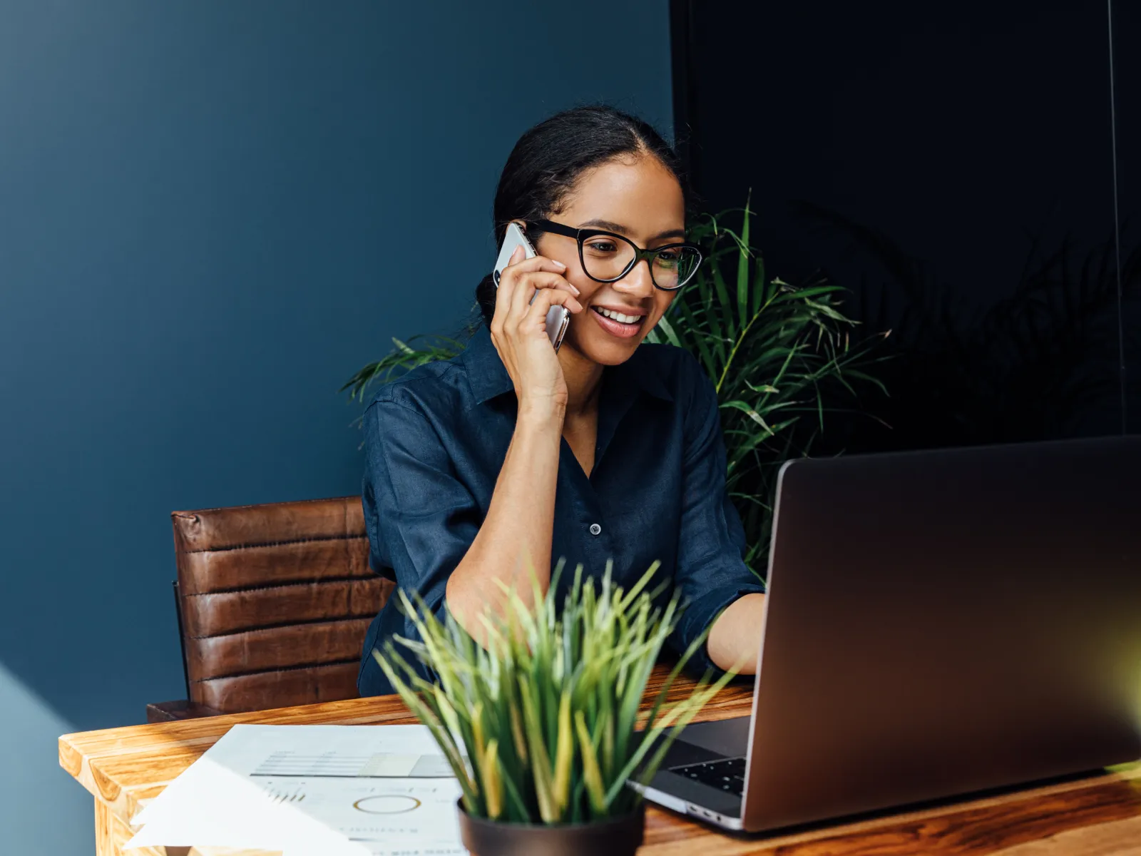 Young professional woman smiling while talking on phone and working on laptop at a wooden desk