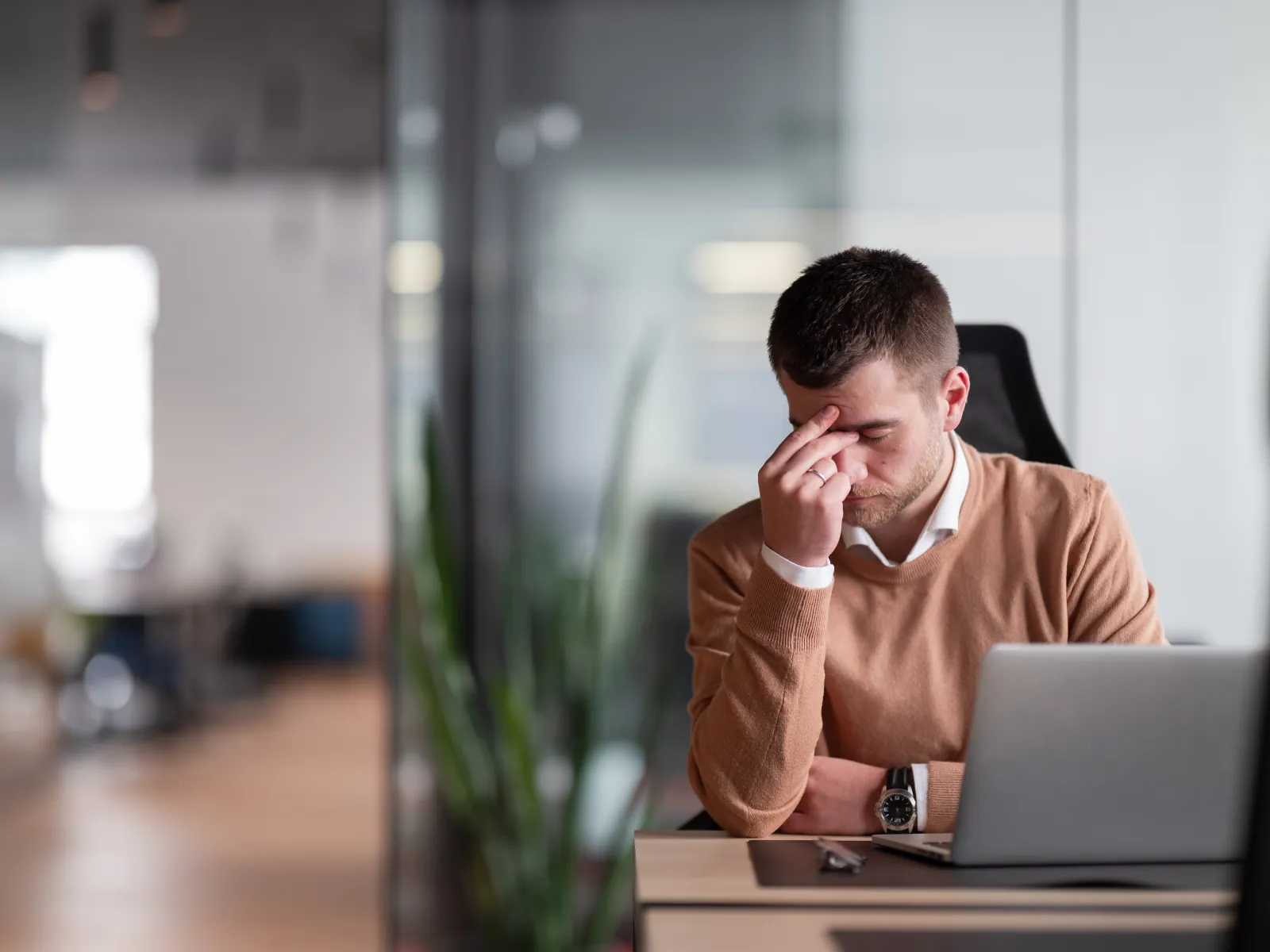 Stressed young man in brown sweater sitting at office desk with laptop, rubbing forehead in frustration.