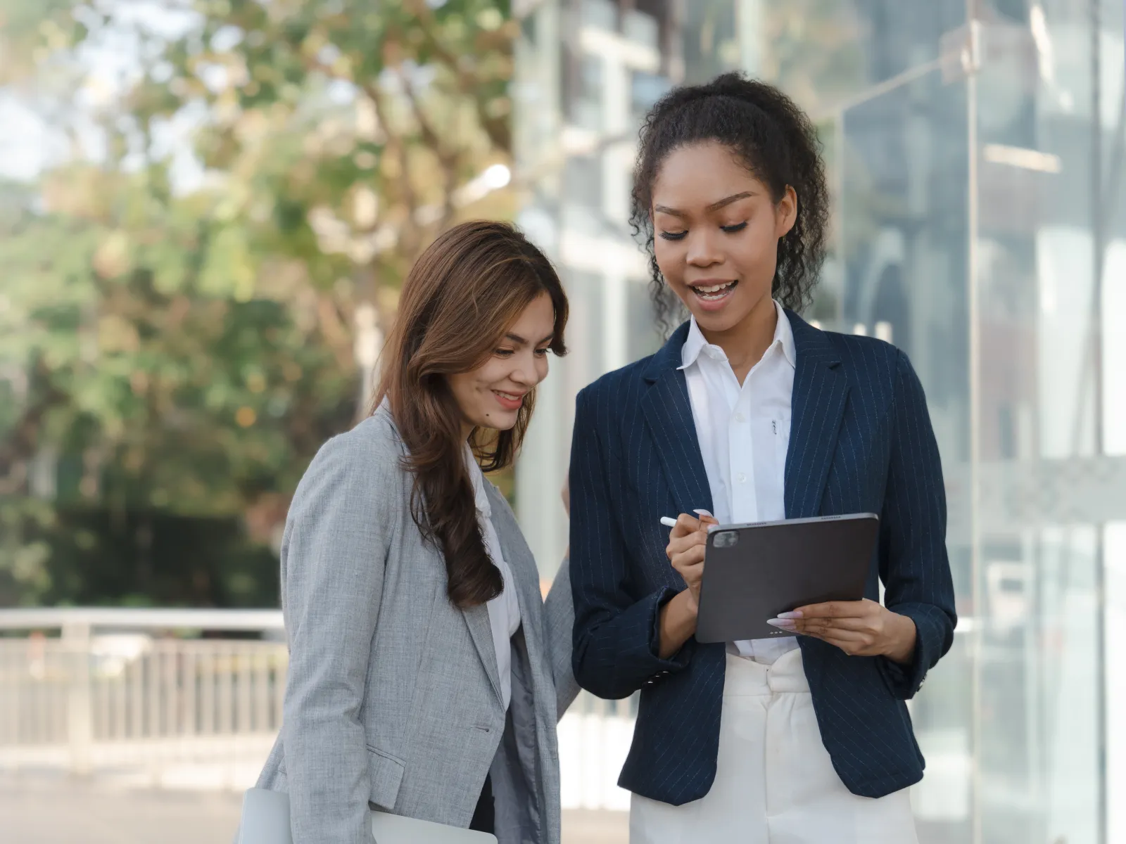 Two professional women discussing notes on a digital tablet outdoors near a modern building.