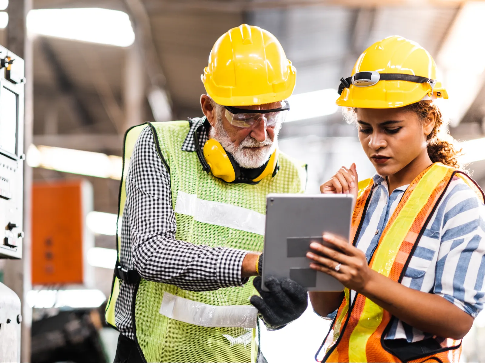 Two factory workers in safety gear using a tablet for checking machinery in an industrial setting