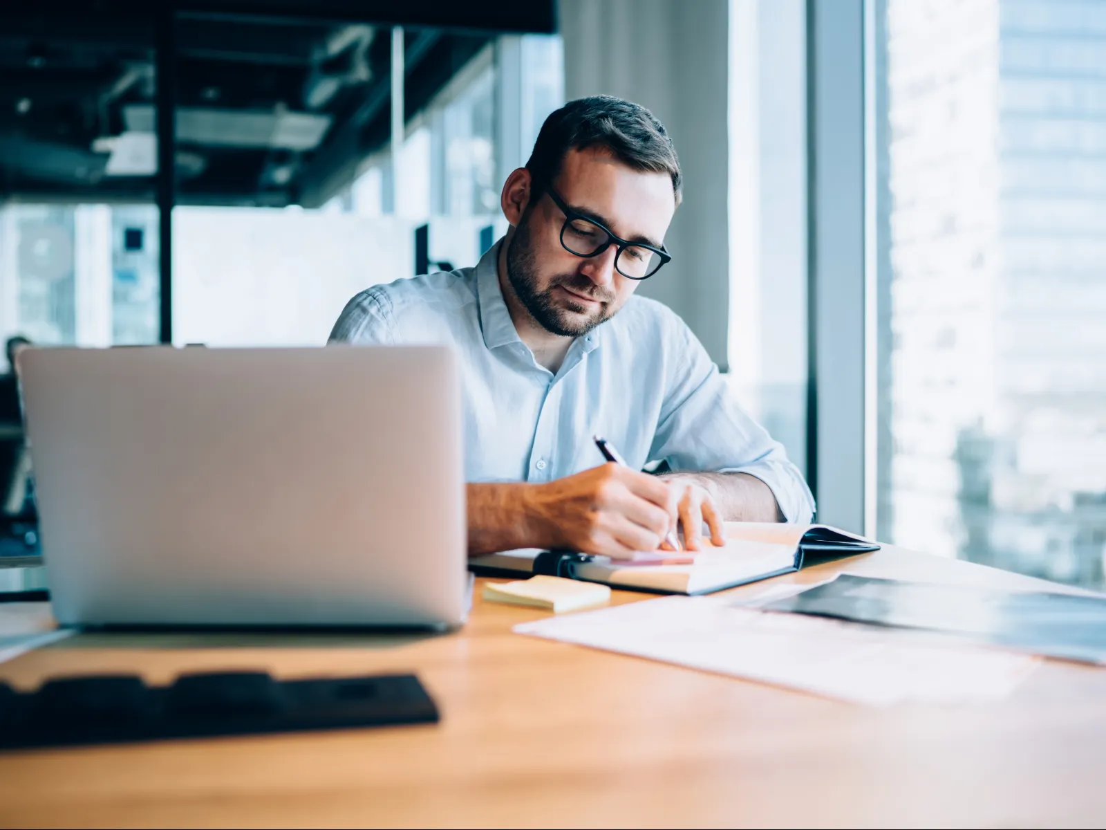 Man with glasses working on notes at desk with laptop and documents in modern office environment