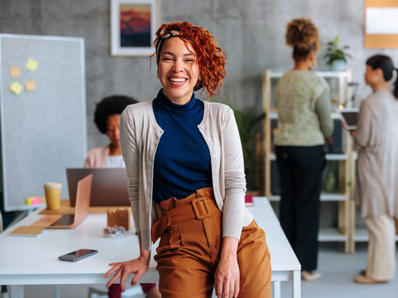 Smiling woman with curly red hair wearing a cardigan and high-waisted pants in a modern office setting.