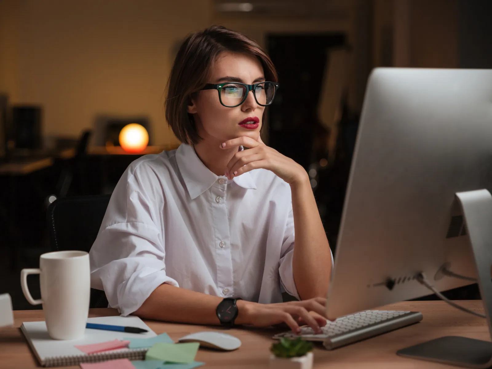 Young woman wearing glasses working thoughtfully at a computer in a cozy, dimly lit office environment.