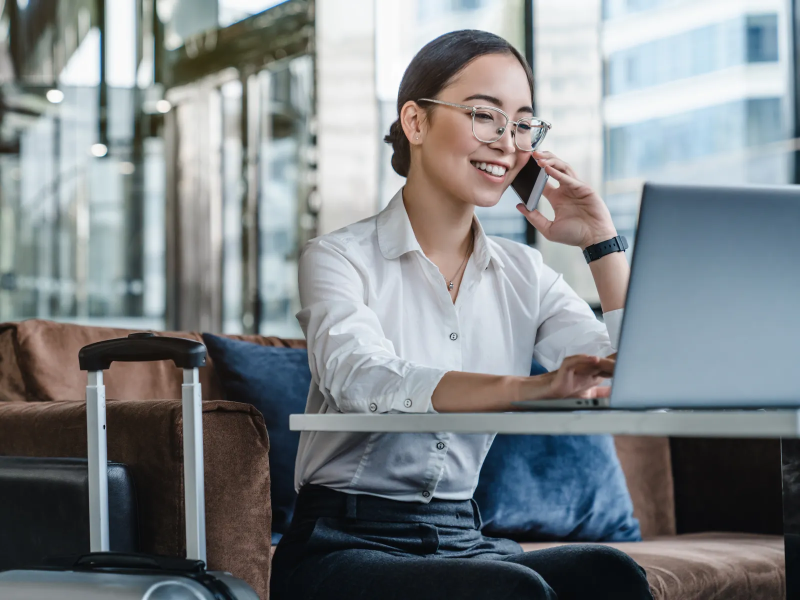 Young businesswoman with glasses uses laptop and talks on phone in modern airport lounge with suitcase nearby