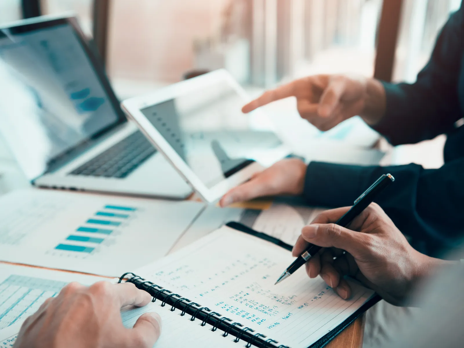Two people analyzing data on a tablet and writing notes in a notebook near a laptop with charts on the table.