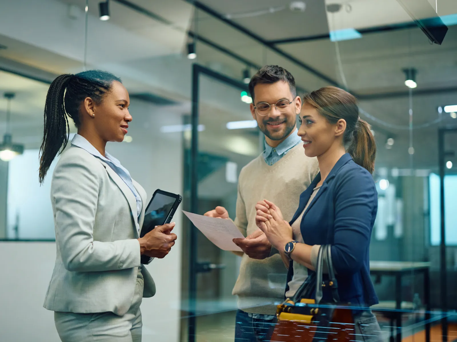 Three professionals engaged in a discussion inside a modern office with glass walls, holding documents and a tablet.