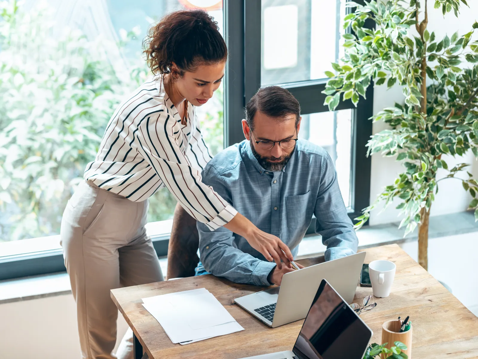 Woman in striped shirt helps man in blue shirt with laptop work in bright office surrounded by plants.