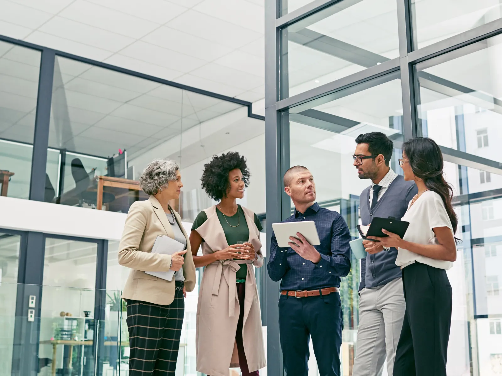 Five diverse professionals standing and discussing with digital tablets in modern bright office space.