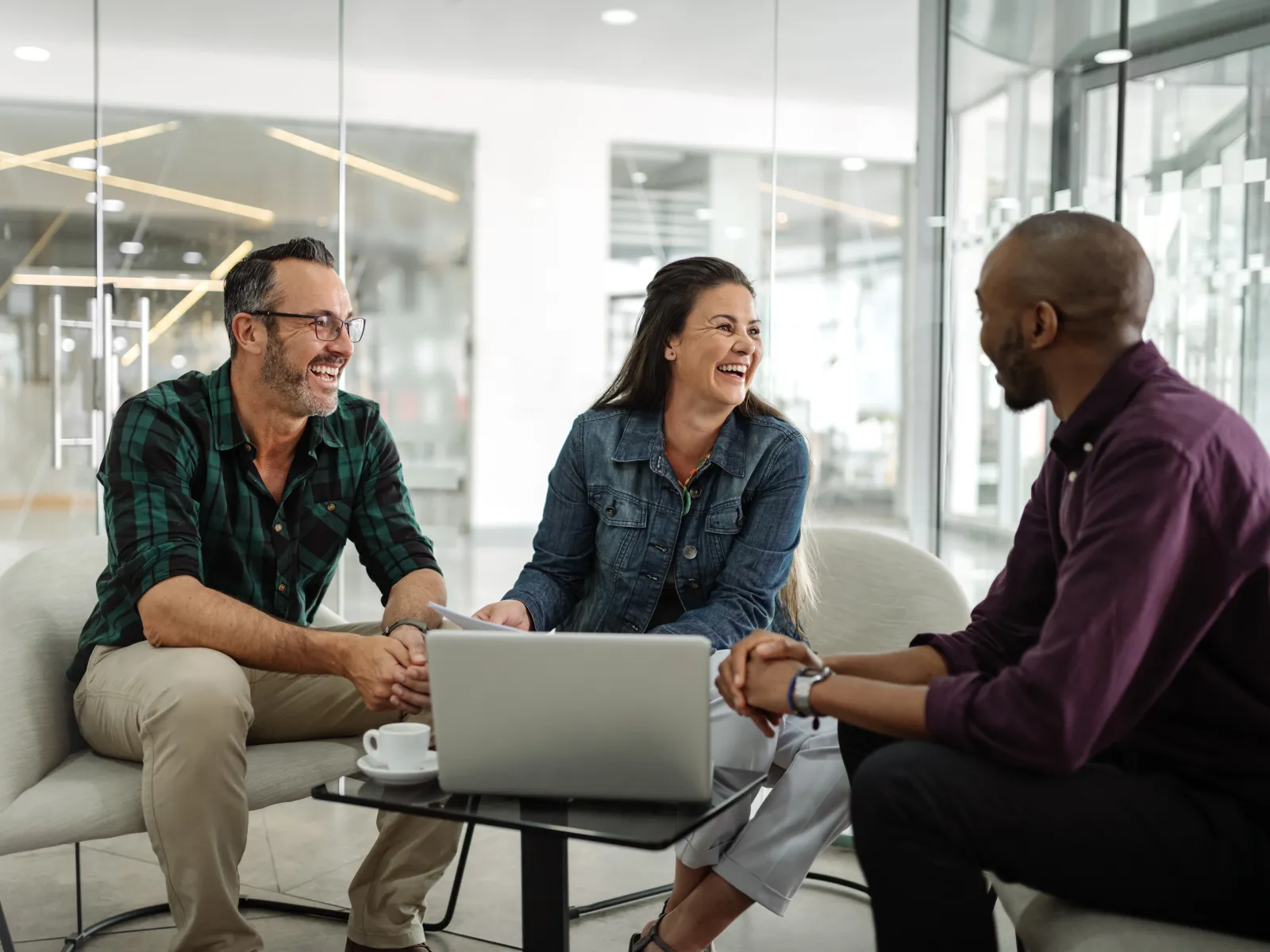Three colleagues smiling and discussing work around a laptop in a modern office lounge area.