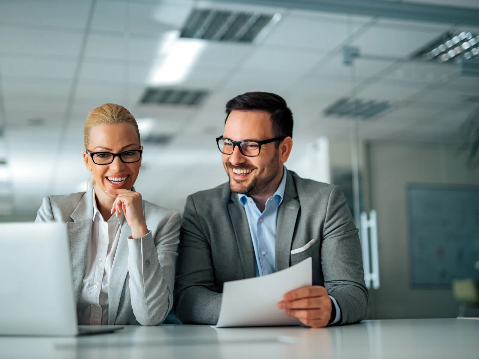 Two business professionals in glasses smiling and discussing documents in modern office setting.