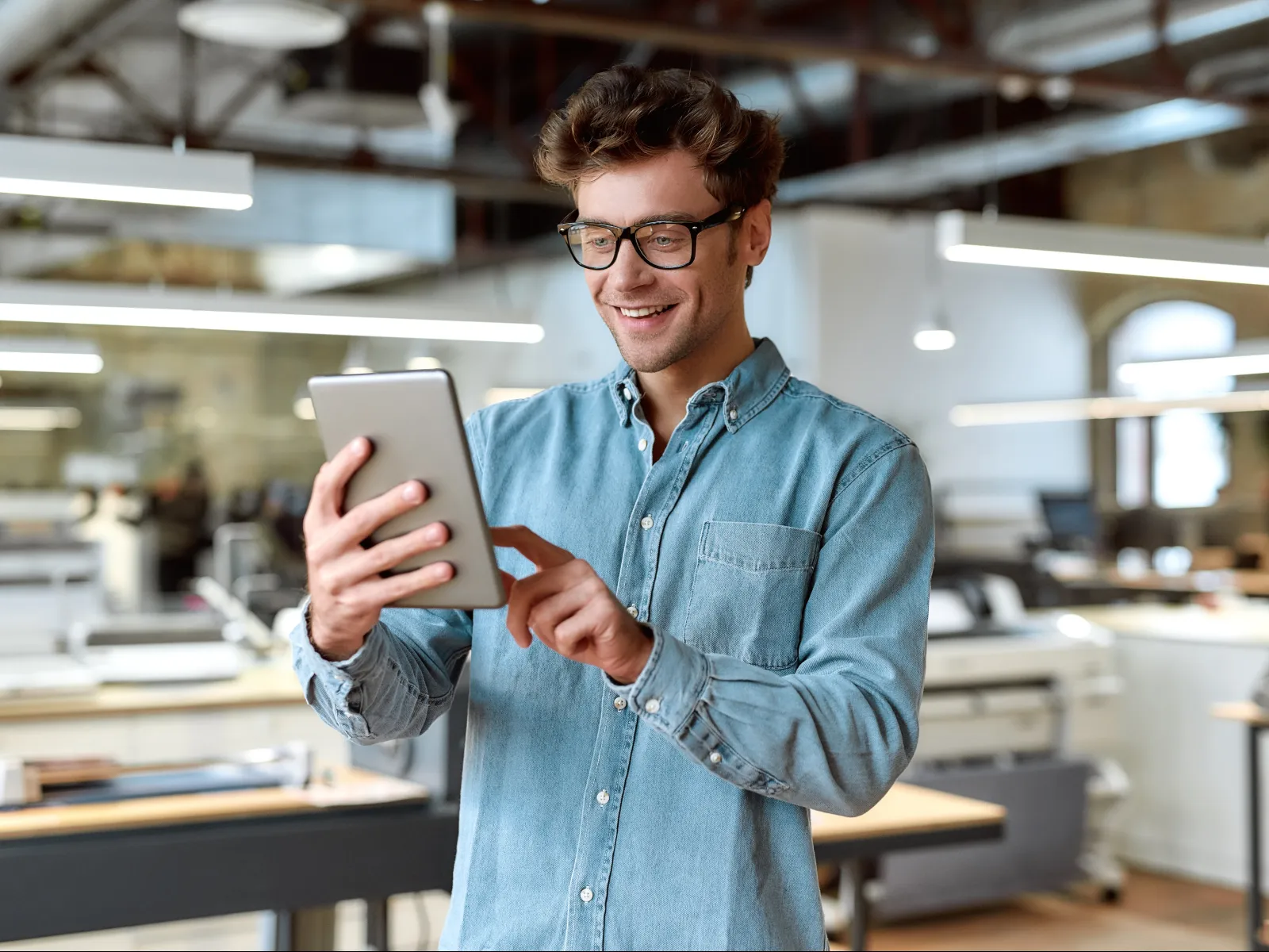 Smiling man in glasses using a tablet in a modern open office space with bright lighting.