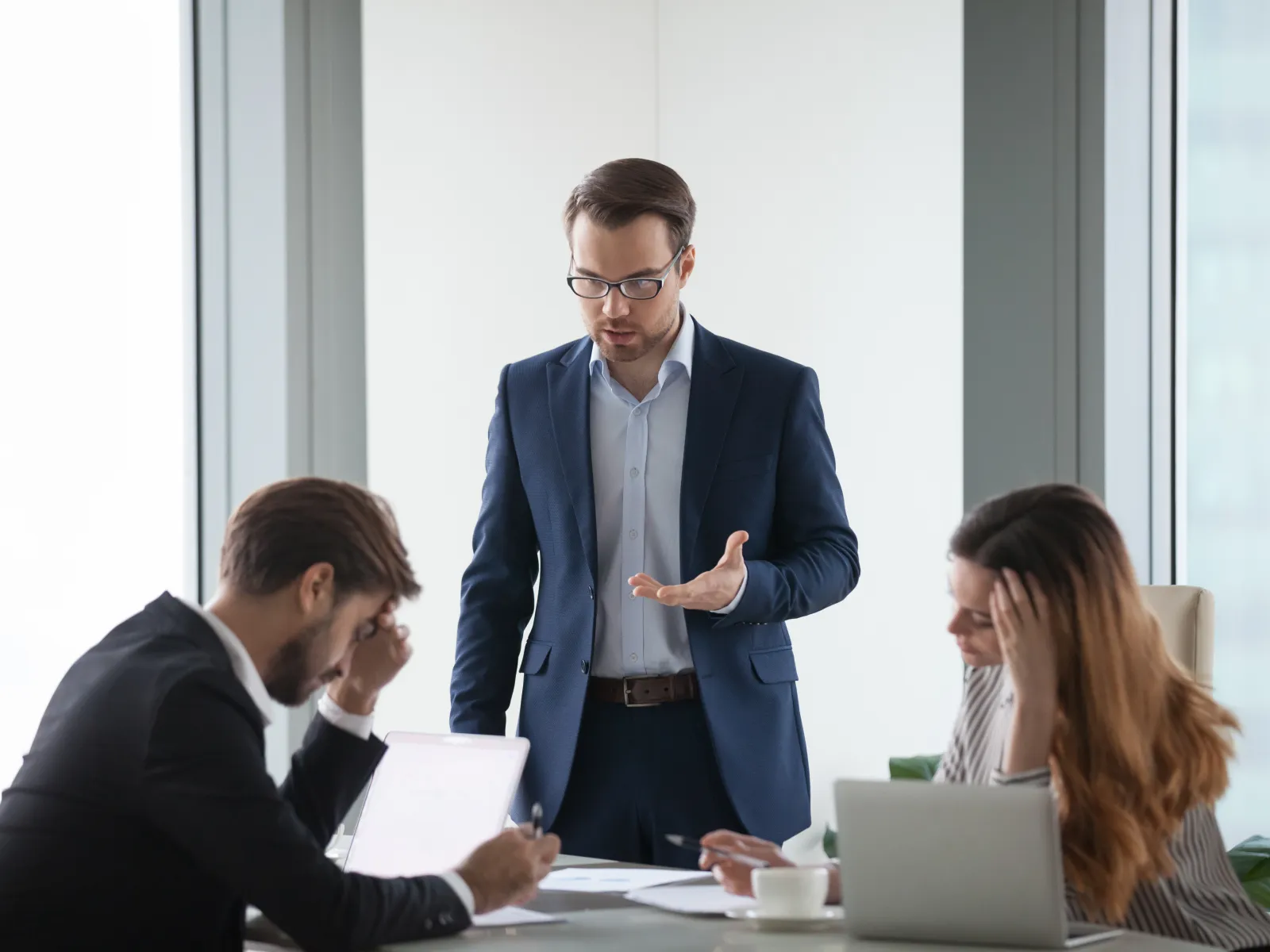 Businessman in suit discusses project with stressed colleagues in modern office meeting room.