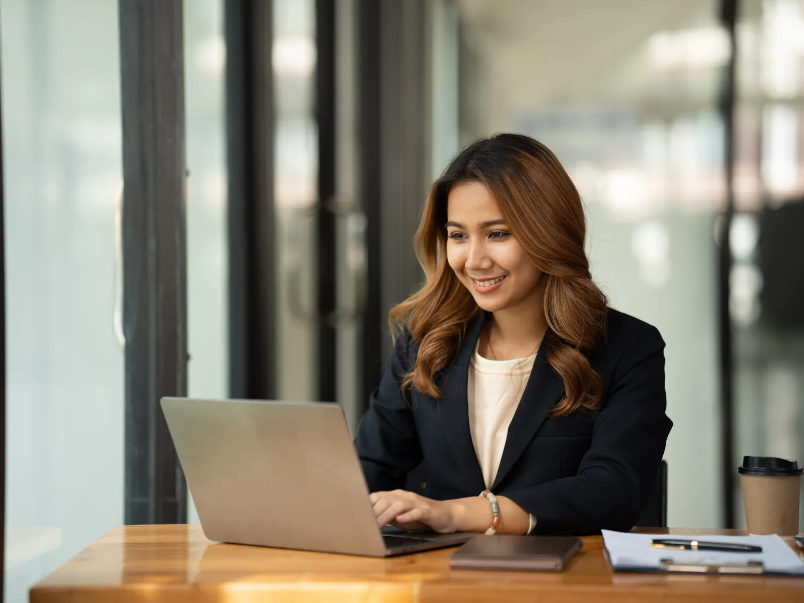 Young professional woman working on laptop in modern office with coffee and documents nearby.