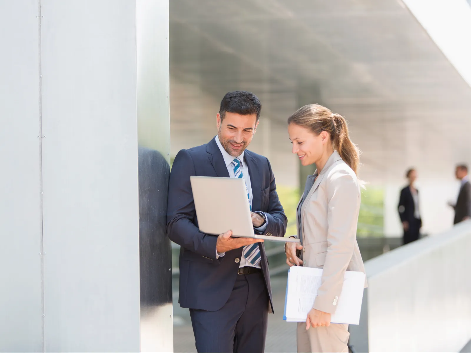 Businessman and businesswoman discussing work on a laptop outdoors near modern office building.