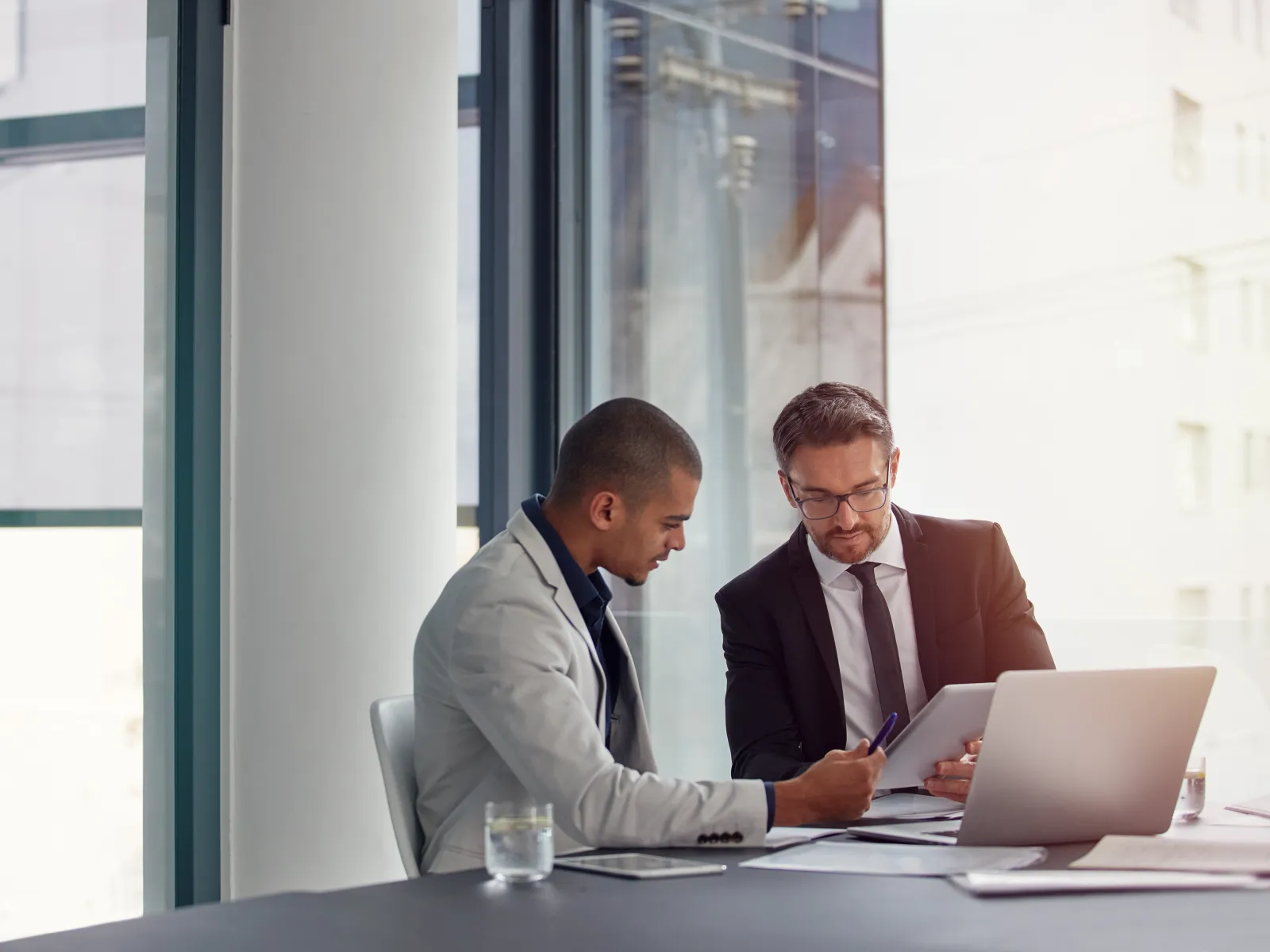 Two businessmen in suits discussing documents in a modern office with a laptop and glass of water on the table