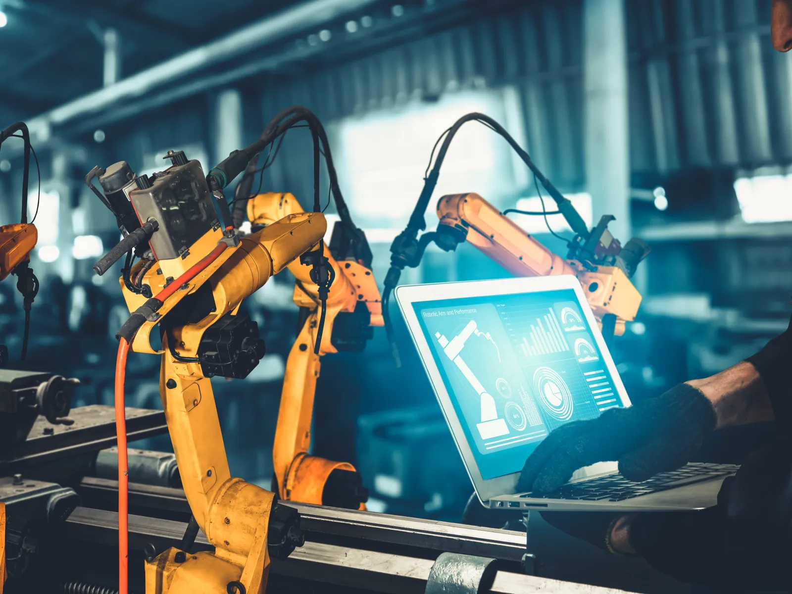 Engineer using laptop to control orange robotic arms in a modern industrial automation facility.