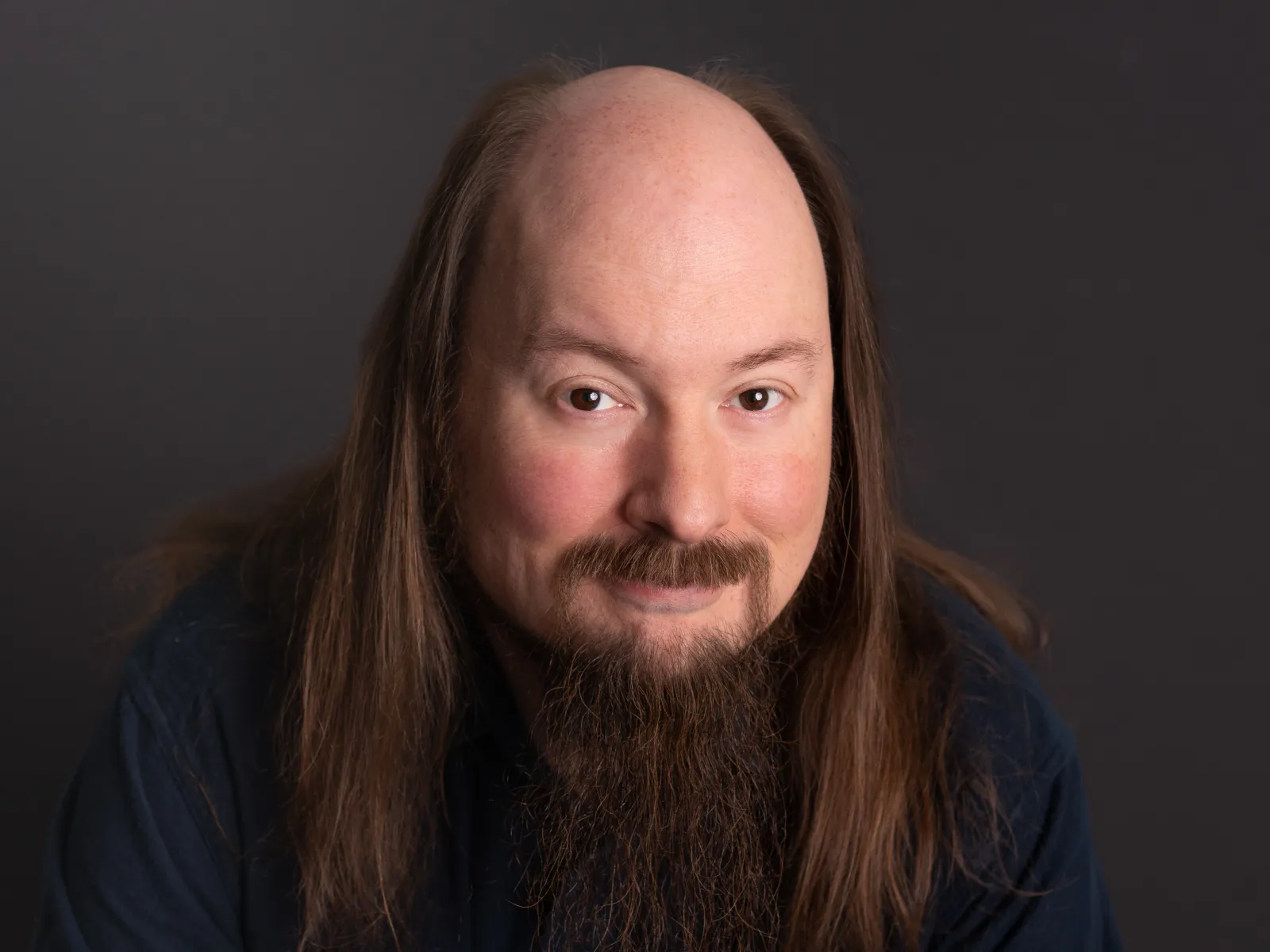 Smiling man with long hair, bald head, and beard leaning on a wooden table with clasped hands