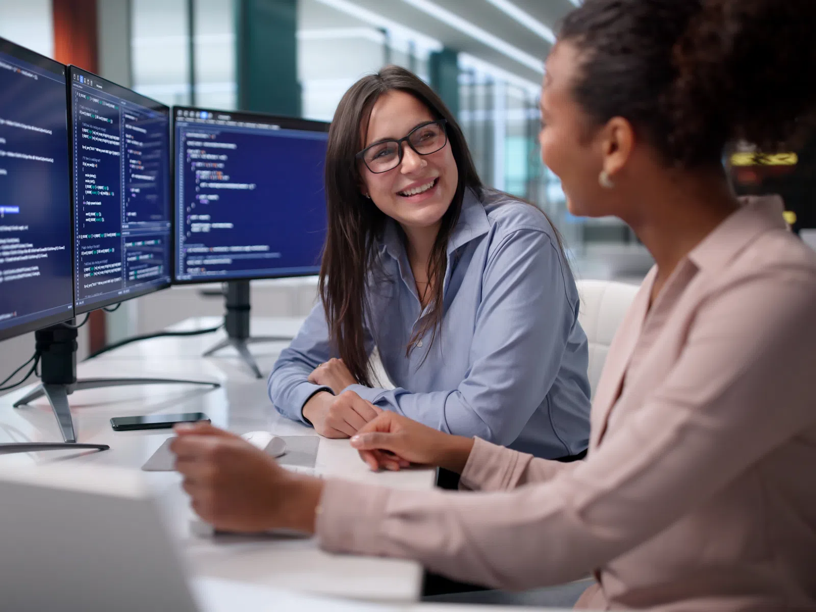 Two female software developers smiling and discussing code on multiple monitors in a modern office environment.