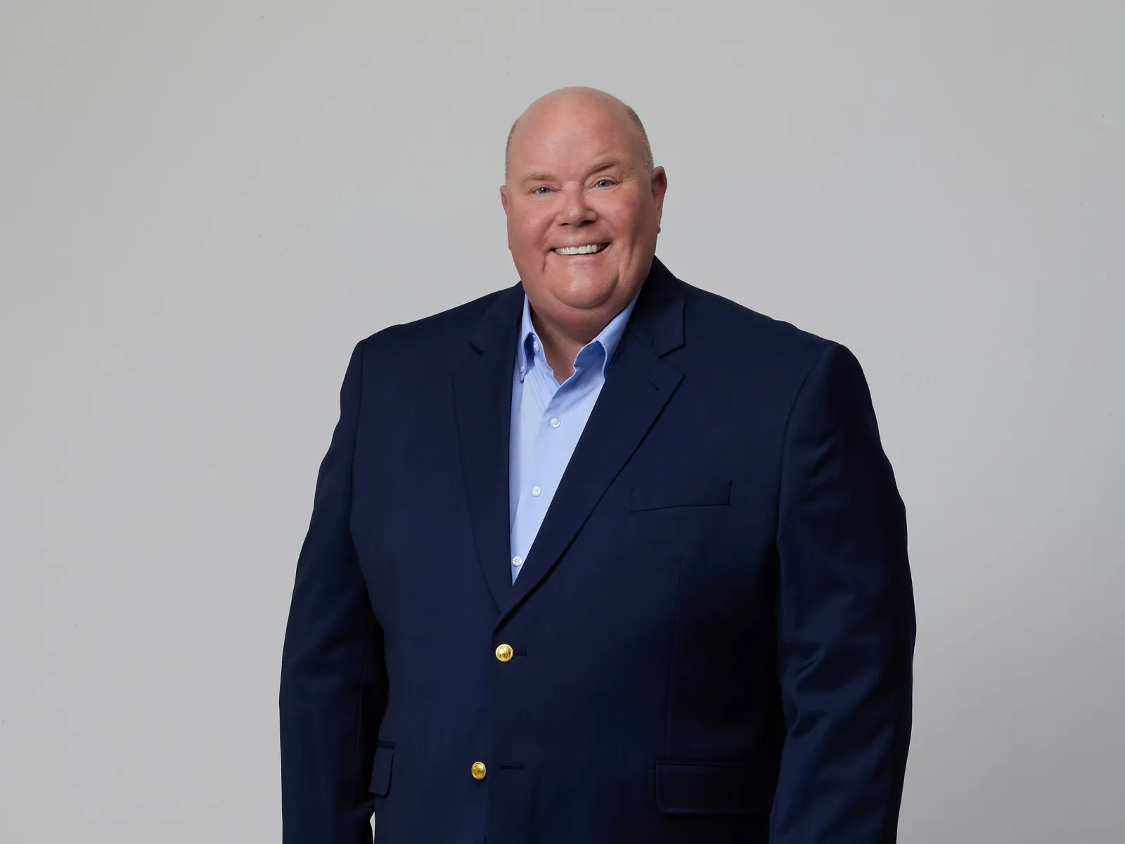 Smiling bald man wearing navy blazer, light blue shirt, khaki pants, and brown shoes standing against gray background.
