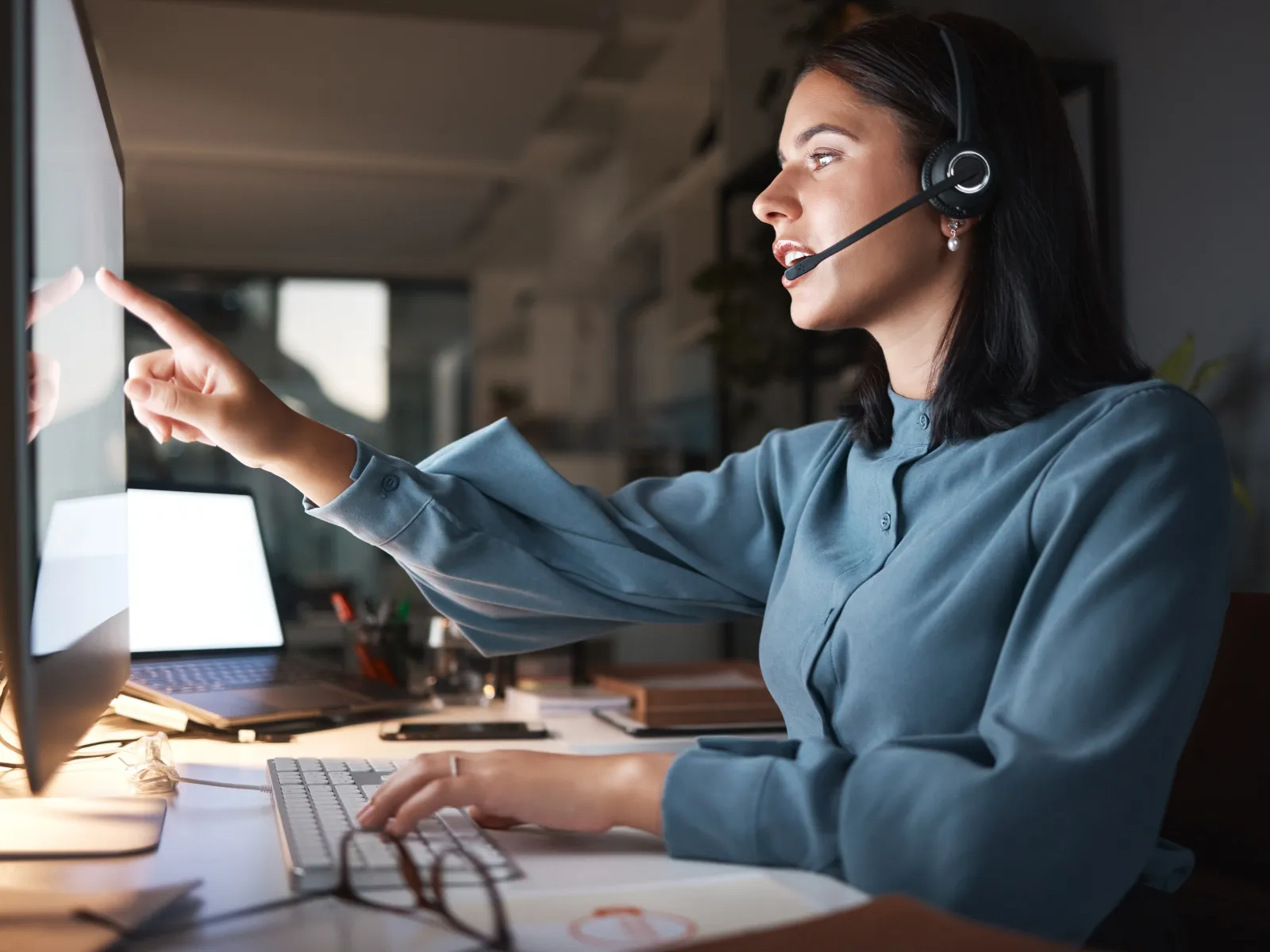 Customer service representative wearing a headset working late on a computer, pointing at the screen in an office.