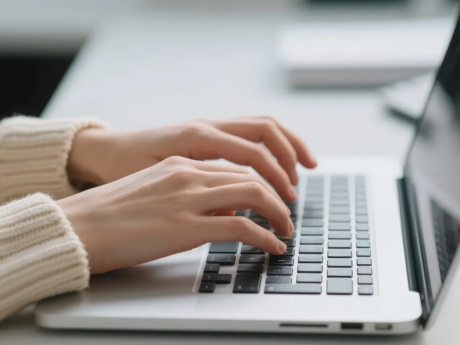 Hands typing on a laptop keyboard in a bright workspace with neutral tones and blurred background.