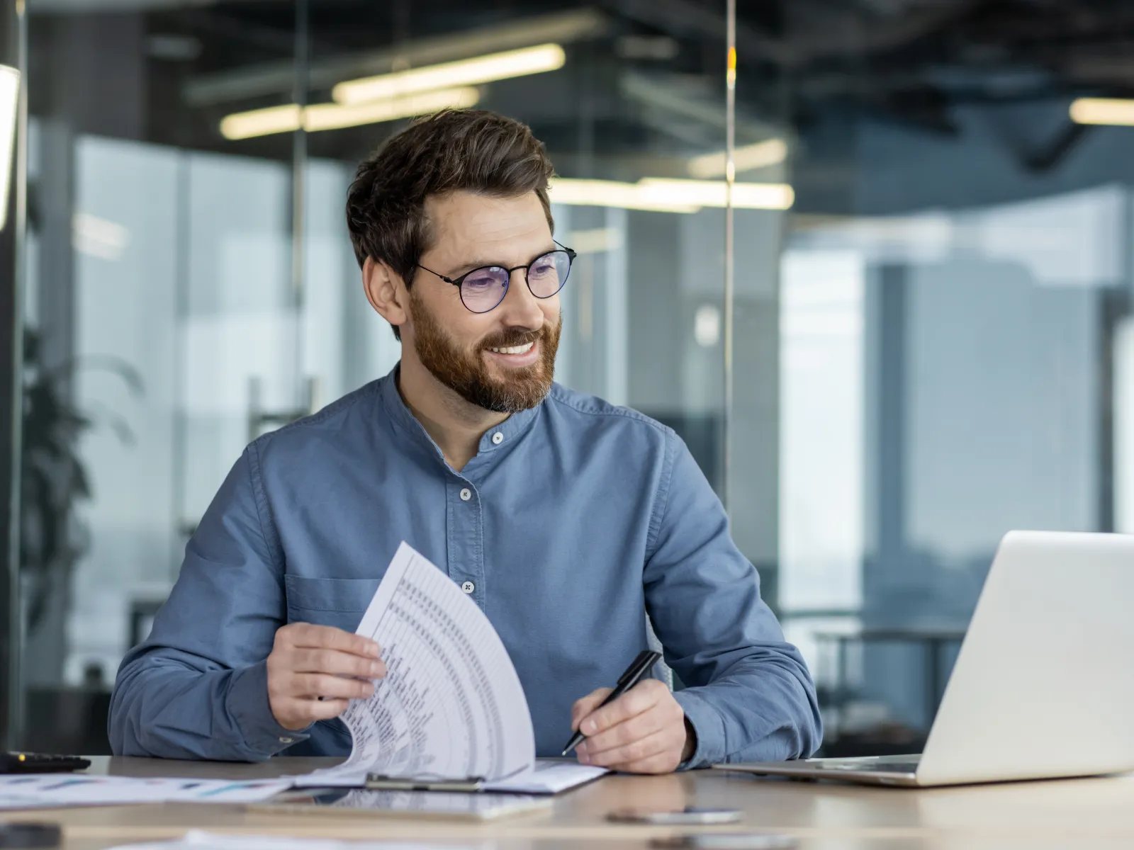 Smiling man in glasses reviewing documents while sitting at office desk with laptop in modern workspace