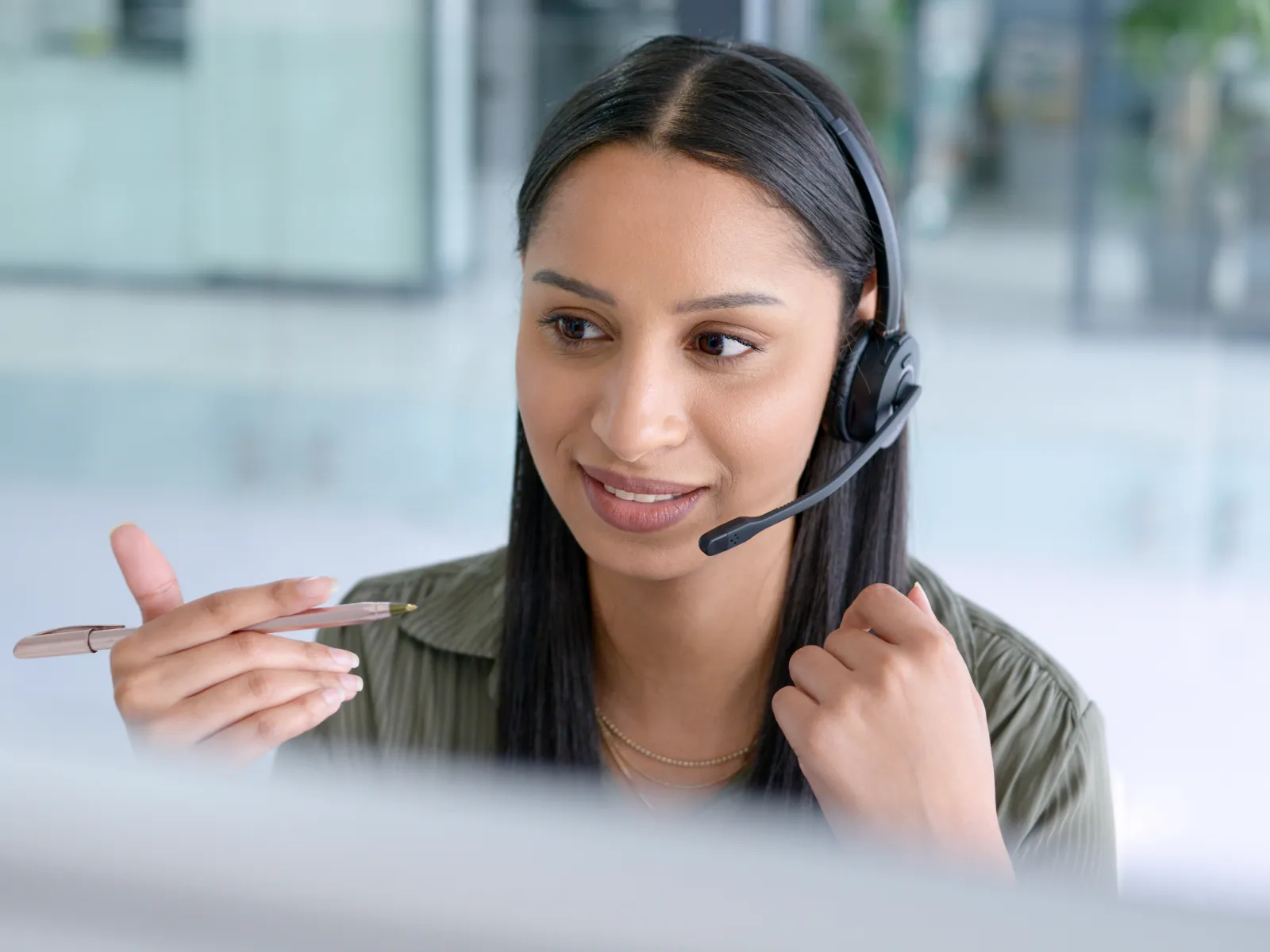 Young woman with headset speaking and pointing with pen during customer support call in office.