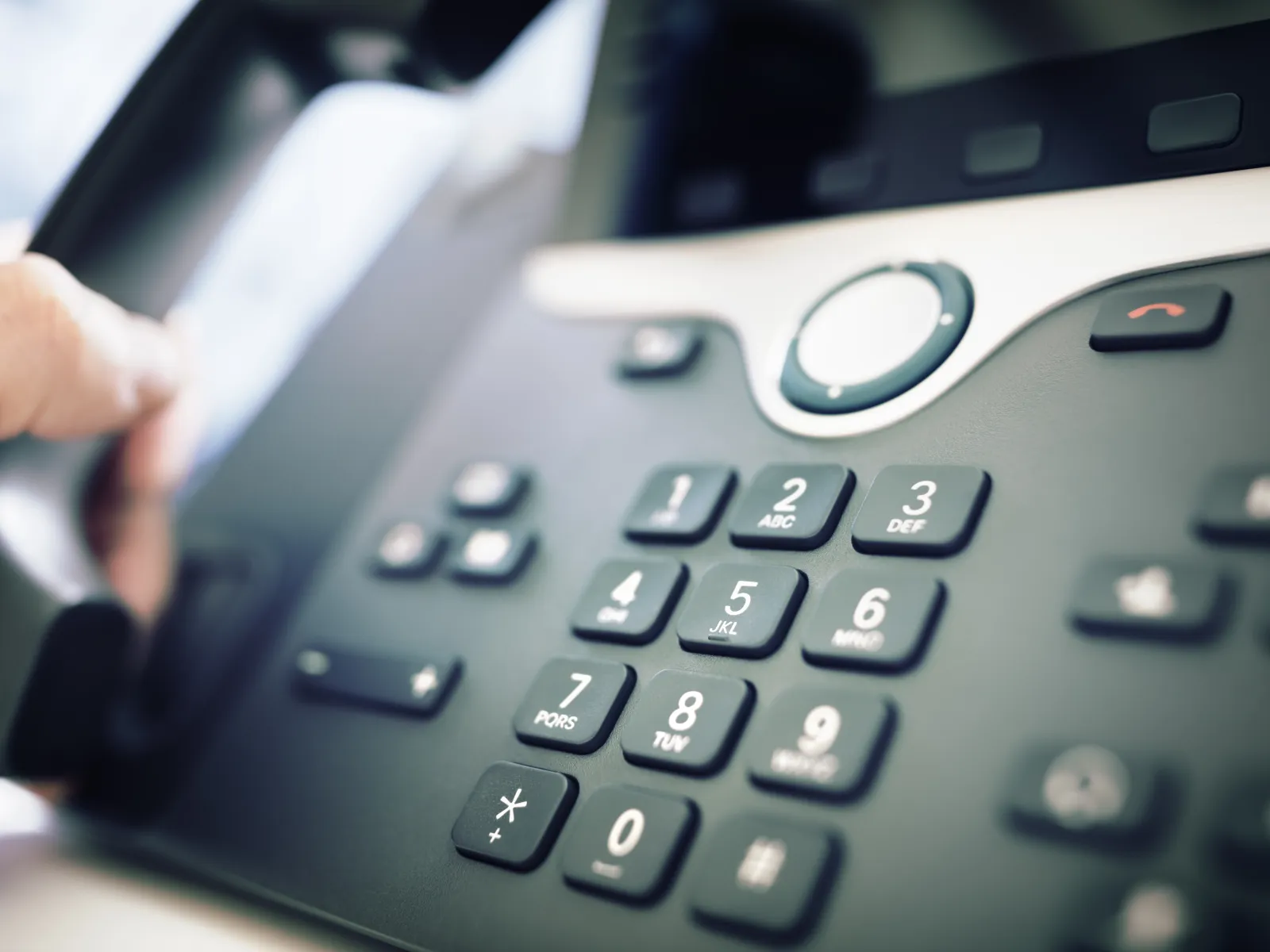 Close-up of a hand holding a phone receiver near a modern office desk phone with numeric keypad.