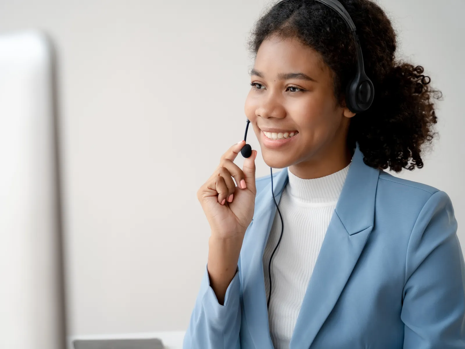 Smiling customer service representative wearing headset and blue blazer talking at computer in modern office